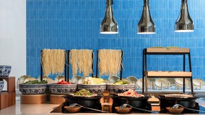 A buffet with various food items and bowls, a wooden stand with a tray, and hanging noodles against a blue tiled wall. - Holiday Inn Express Beijing Tongzhou by IHG (Tongzhou)