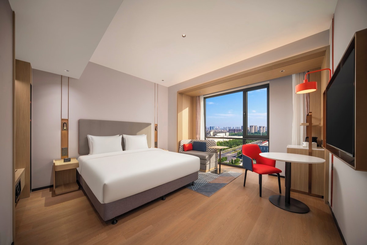 A modern hotel room with a large bed, a desk with a chair, a small table, and a large window offering a city view.