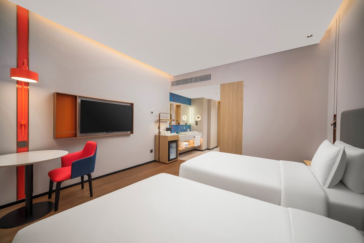 A hotel room with a flat-screen TV, a small desk with a chair, a bed, and a bathroom area.