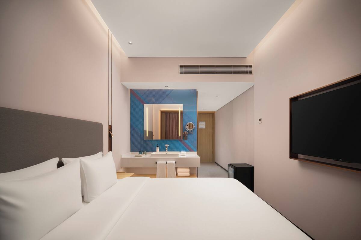 A modern hotel room with a large bed, a bathroom with a mirror and sink, and a TV mounted on the wall.