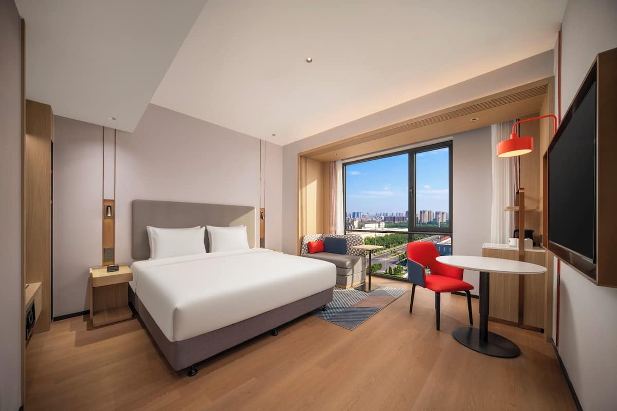 A modern hotel room with a large bed, a desk with a chair, a small table, and a large window offering a city view.
