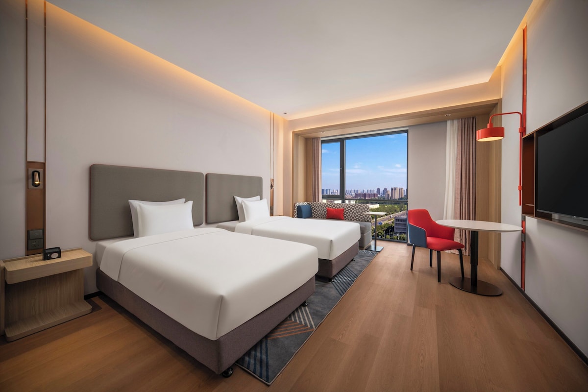 A hotel room with two beds, a TV, a desk, and a view of the city.