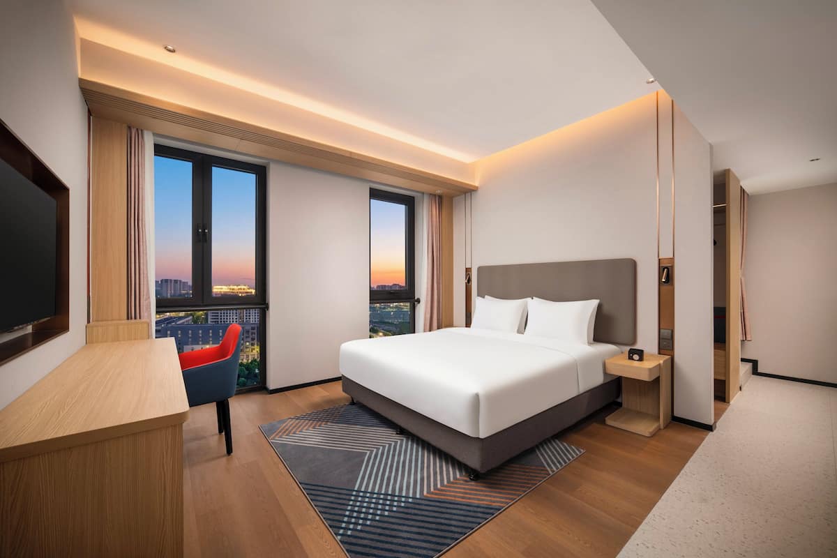 A modern hotel room with a large bed, a desk, a chair, and a view of the cityscape.