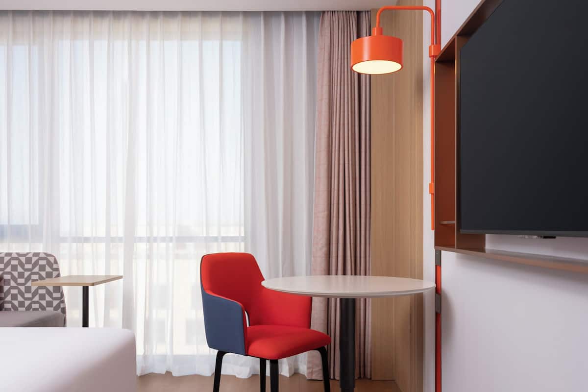A hotel room with a bed, a red and blue chair, a small table, a TV, and a window with sheer curtains.