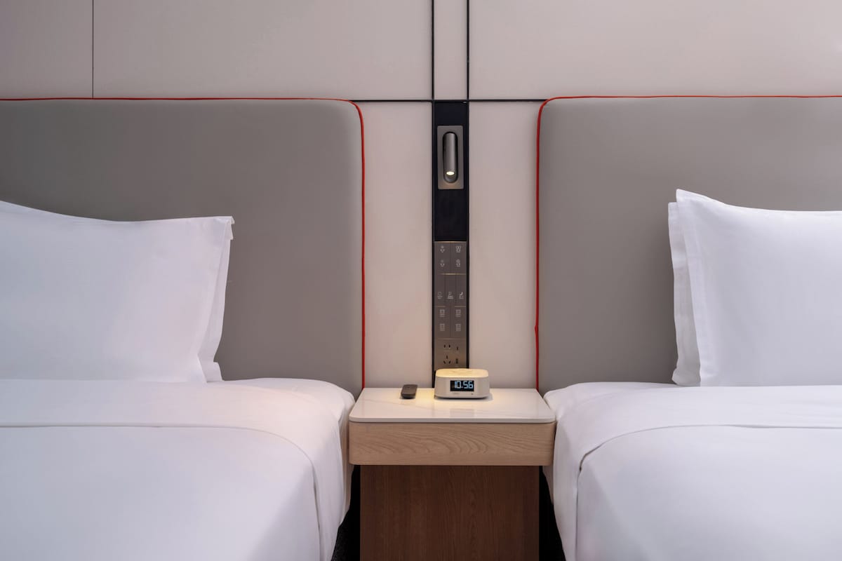 A hotel room with two beds, a nightstand with a clock, and a wall-mounted panel.