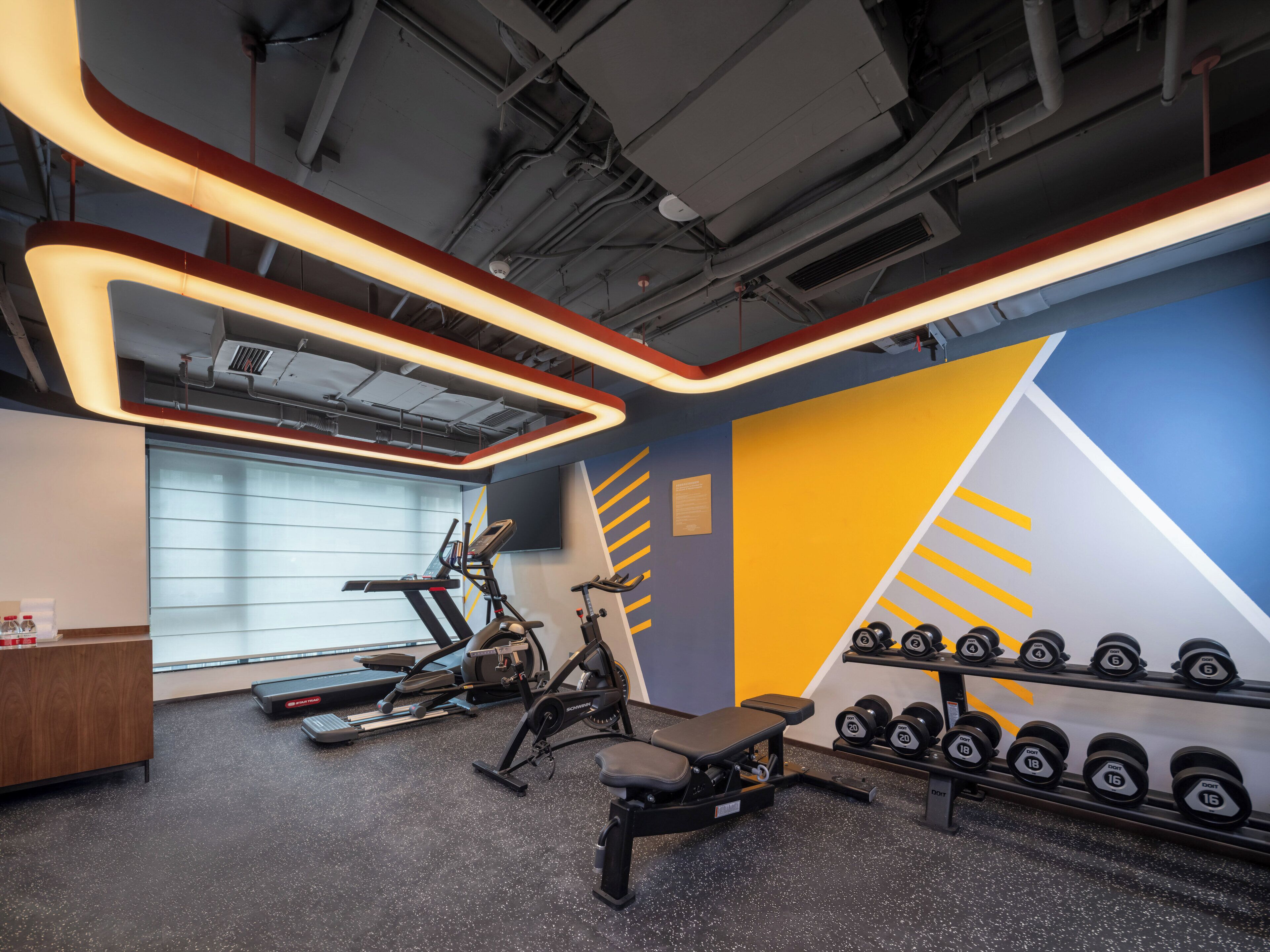 A gym with exercise equipment, a wall with a geometric design, and a row of dumbbells.