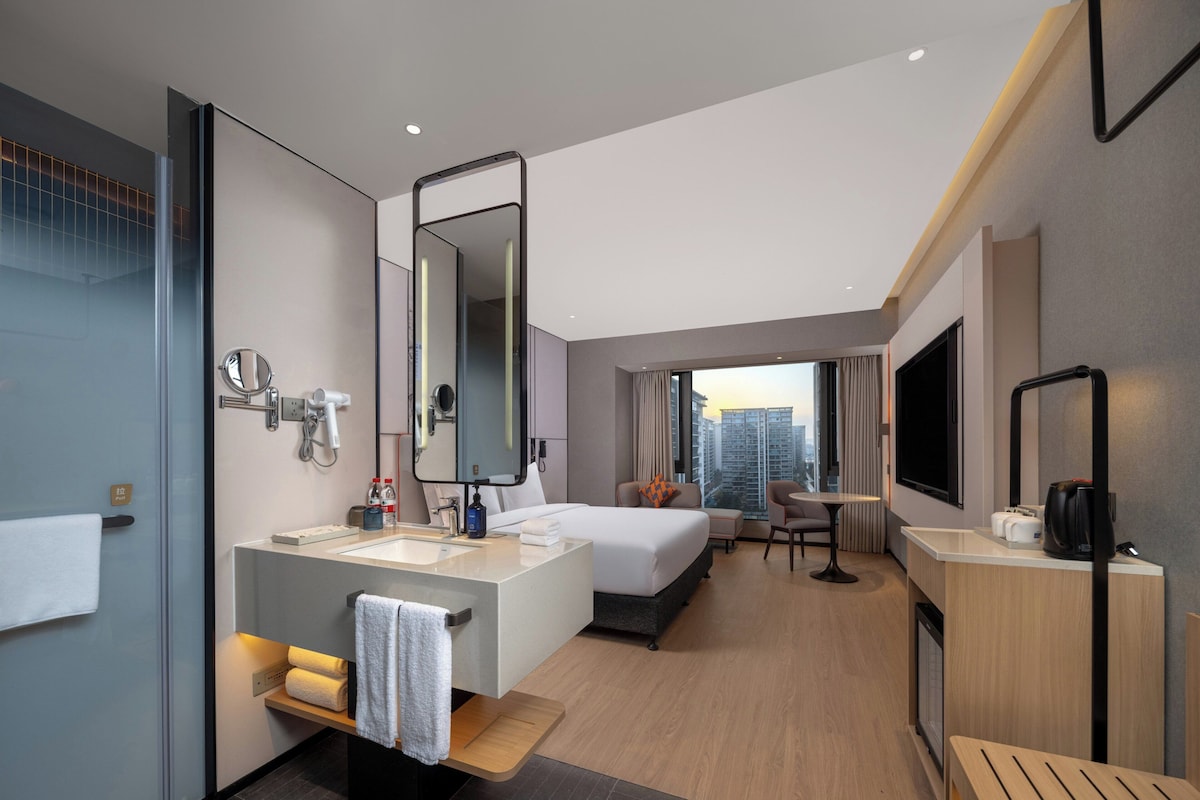 A modern hotel room with a large bed, a sitting area, a bathroom with a vanity, and a city view.