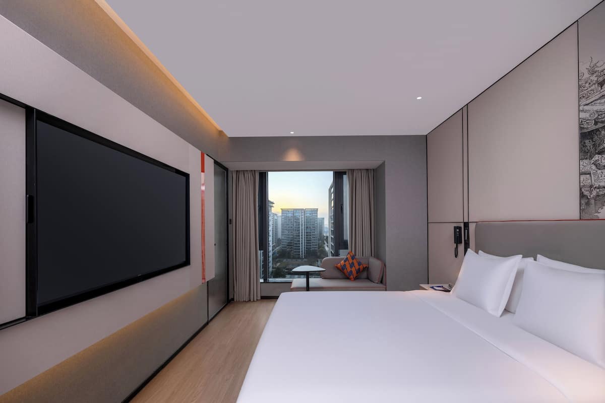 A modern hotel room with a large bed, a flat-screen TV, a small table, and a view of the cityscape.