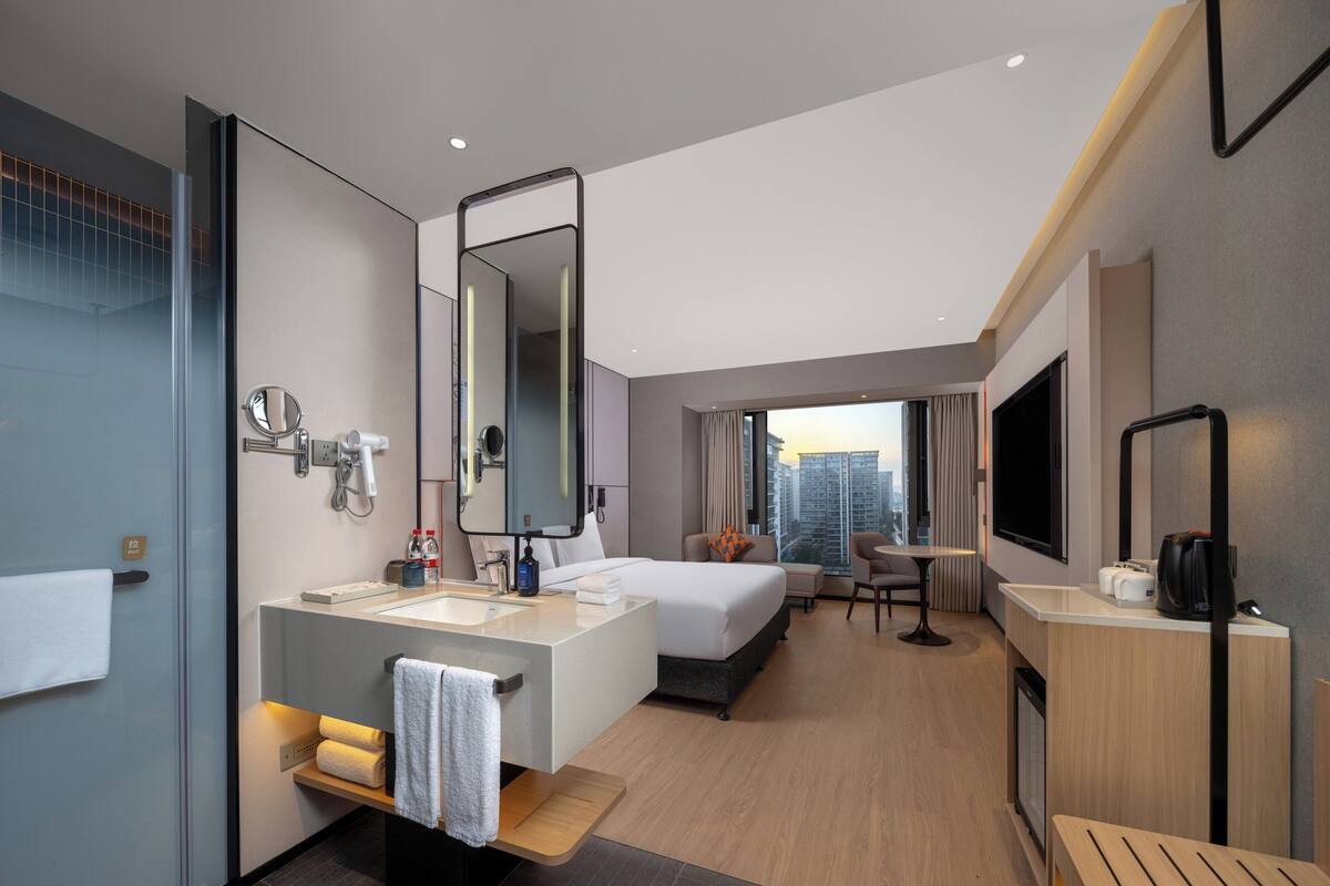 A modern hotel room with a large bed, a sitting area, a bathroom with a vanity, and a city view.