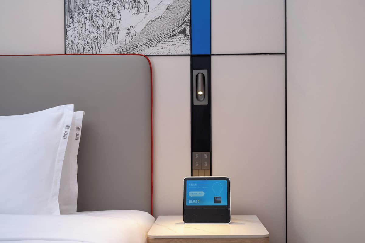 A hotel room with a bed, a nightstand with a digital clock, and a wall-mounted elevator.
