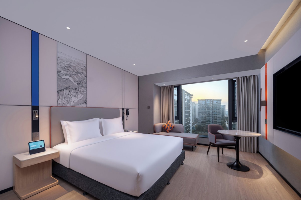A modern hotel room with a large bed, bedside table, and a small desk with a chair.