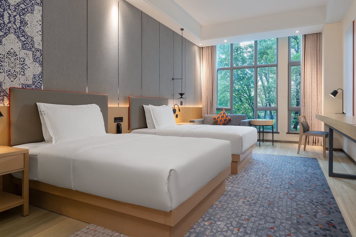A hotel room with two beds, a desk, a chair, a TV, and large windows.