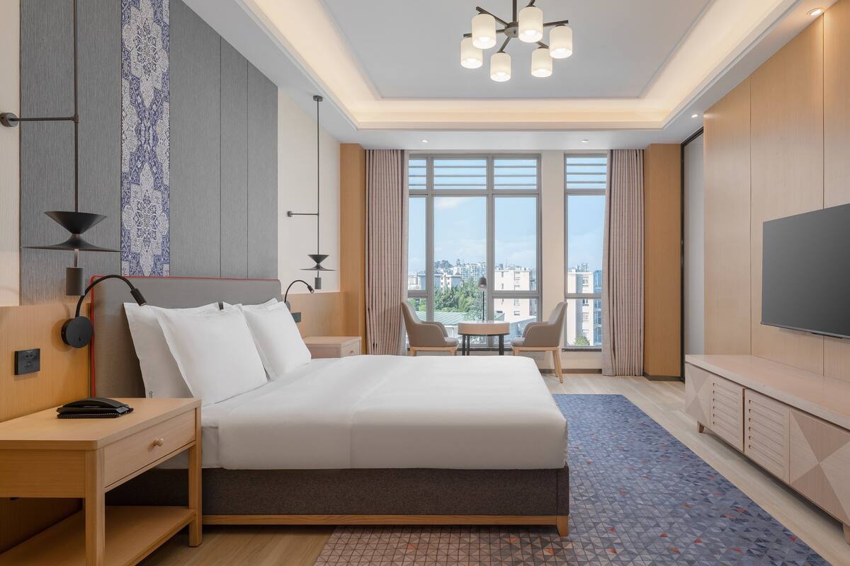 A modern hotel room with a large bed, bedside tables, a desk, a TV, and a view of the city through large windows.