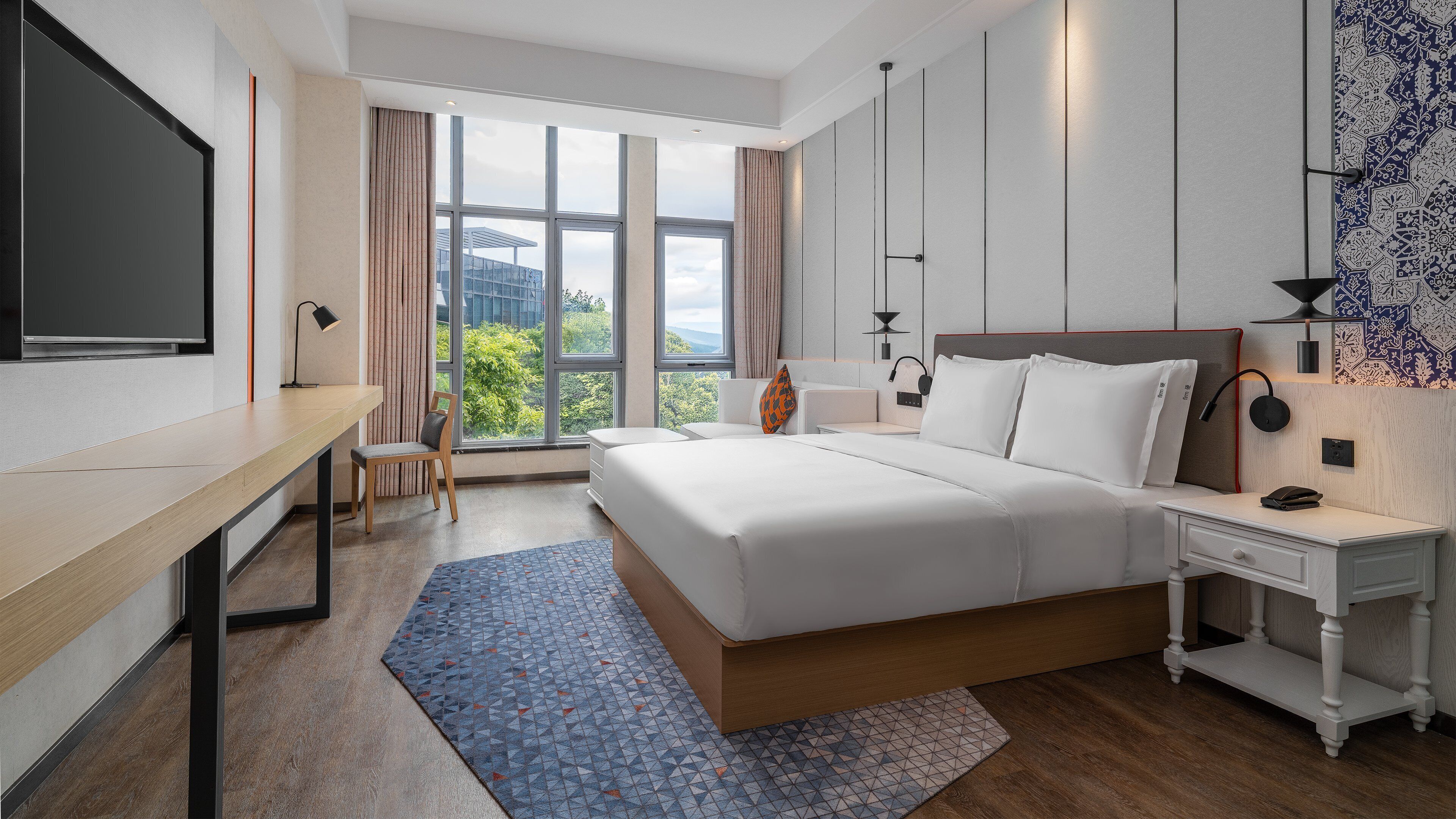 A hotel room with a bed, desk, chair, bedside tables, and a large window with a view.