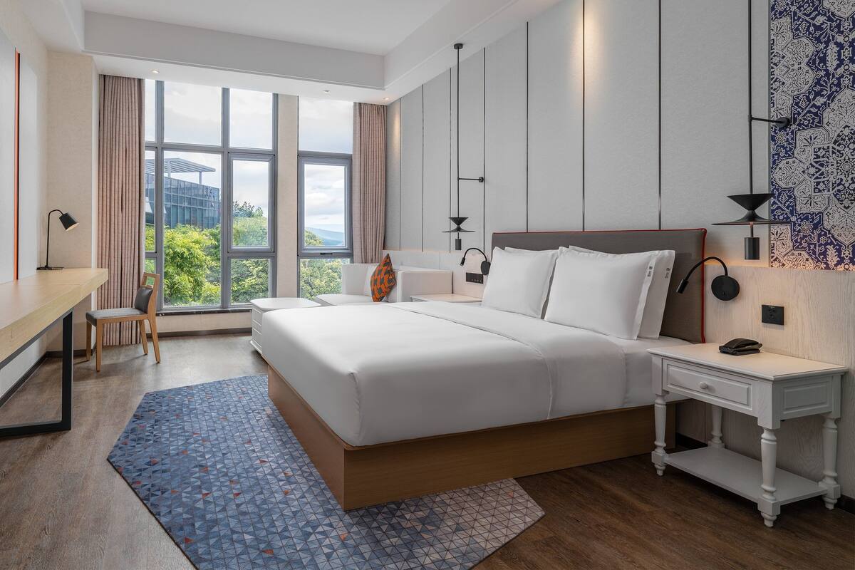 A hotel room with a bed, desk, chair, bedside tables, and a large window with a view.