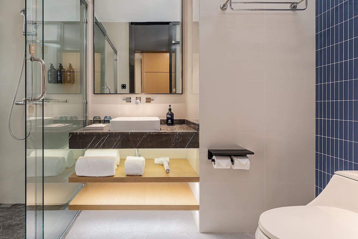 A modern bathroom with a glass-enclosed shower, a sink with a marble countertop, and a wooden shelf with towels.