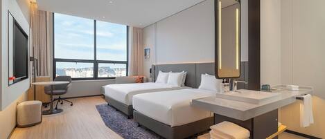 A hotel room with two beds, a desk, a chair, a large window, a bathroom with a mirror, and a view of the city.