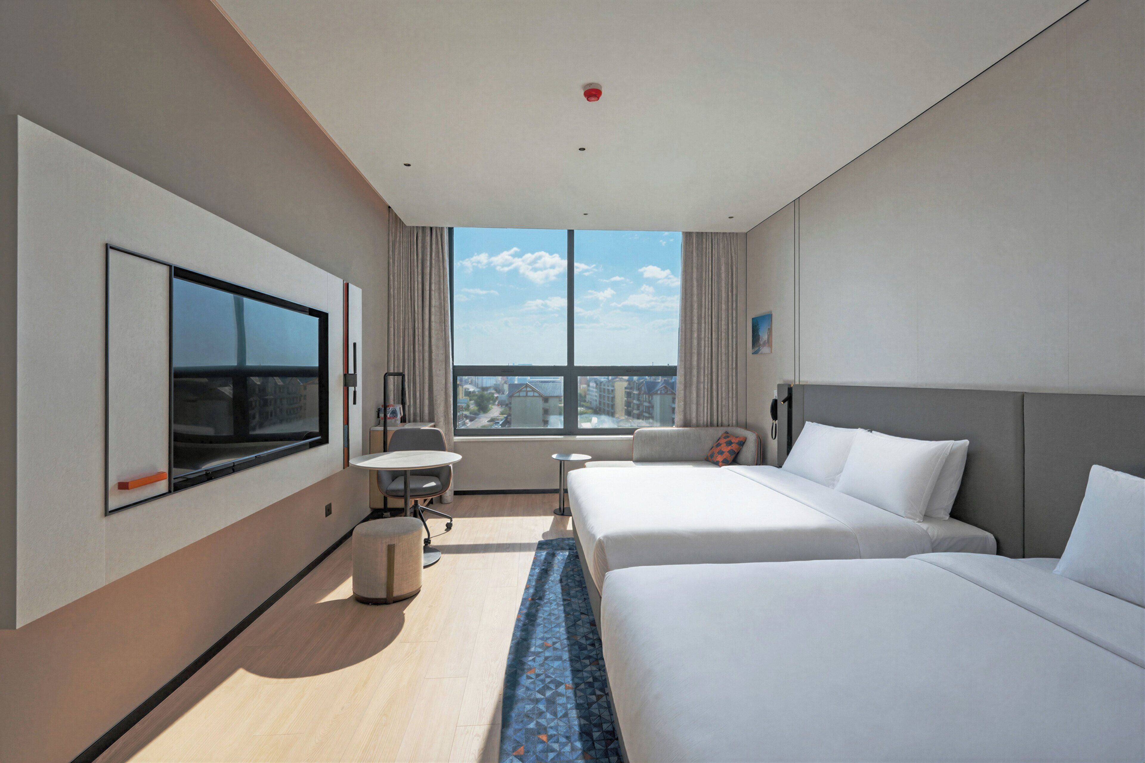 A hotel room with two beds, a TV, a desk, and a window with a city view.