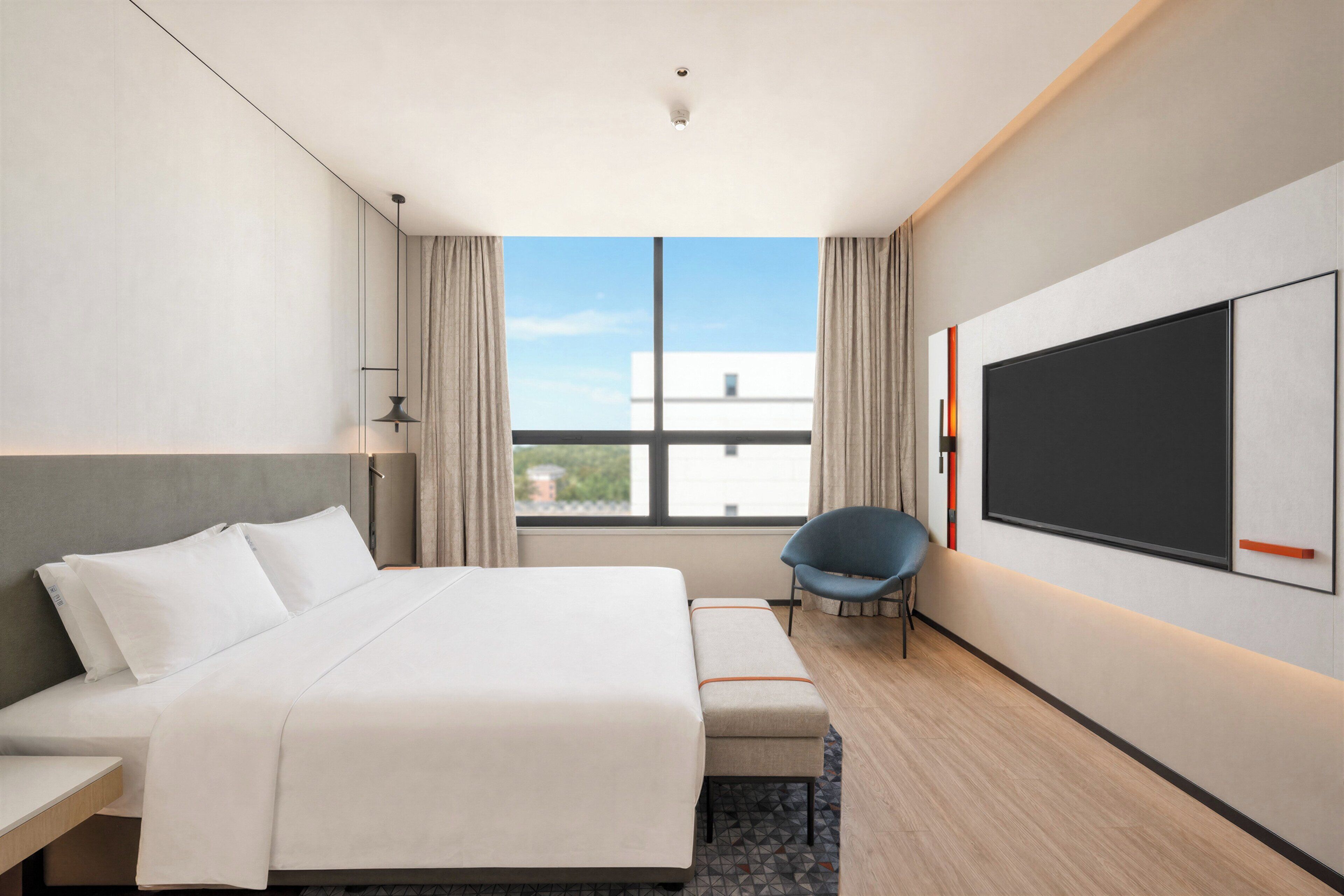 A hotel room with a bed, a chair, a TV, and a window with a view of buildings.