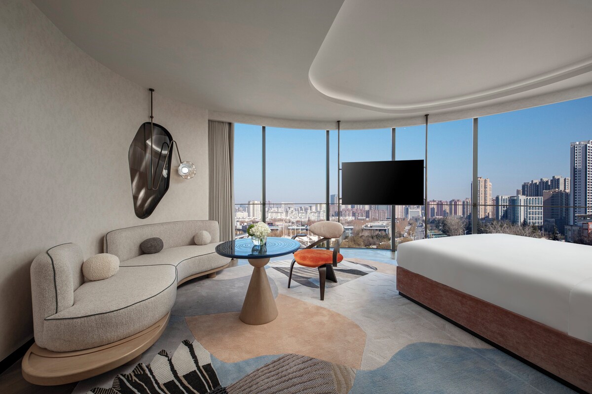 A modern hotel room with a curved sofa, a round coffee table, a bed, and a large TV.