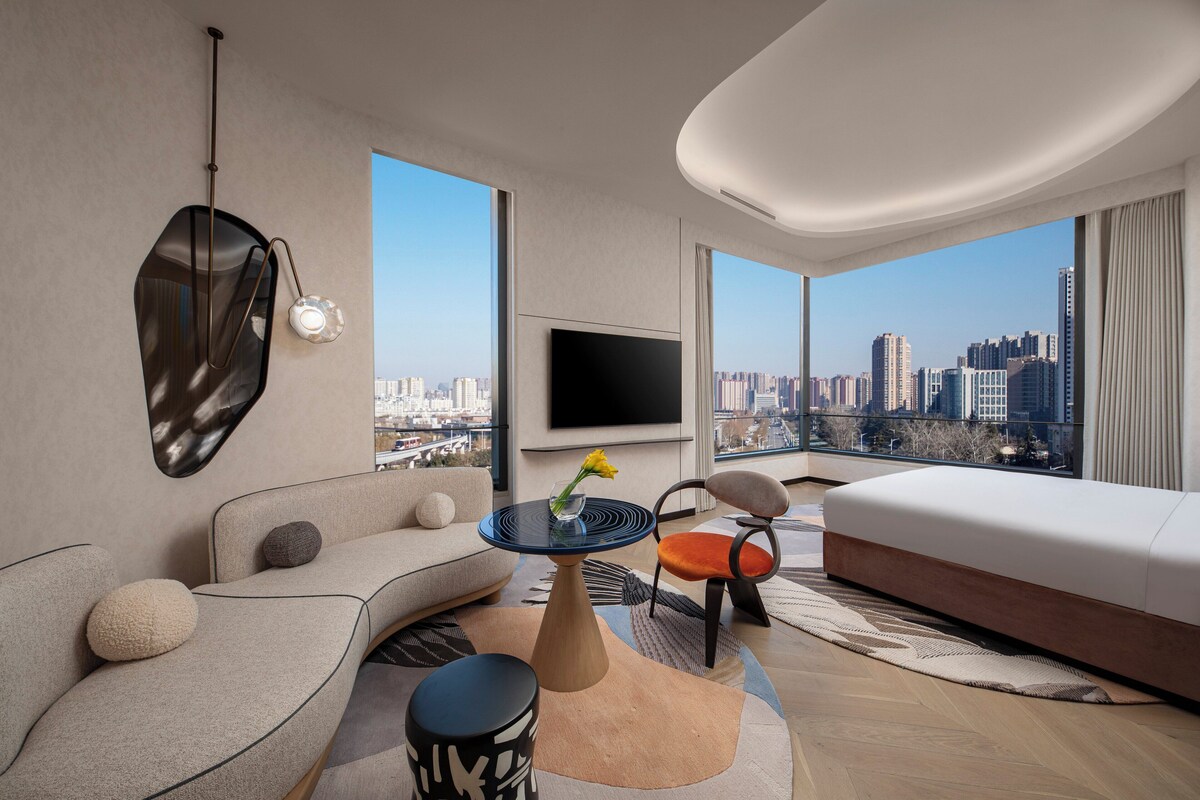 Een moderne hotelkamer met uitzicht over de stad, een flatscreen televisie, een bank, een stoel en een tafeltje met een bloevas.
