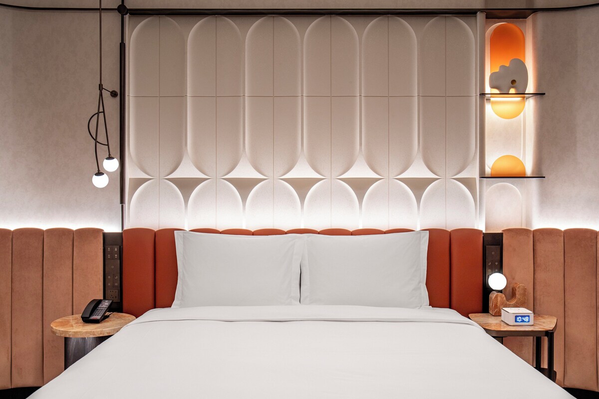 A hotel room with a large bed, a bedside table with a phone, a nightstand with a lamp, and a wall with a decorative panel and a small shelf with a clock.