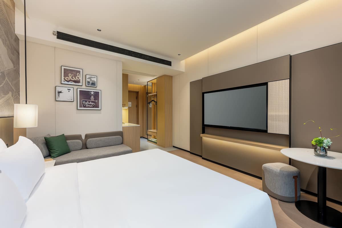 Ein modernes Hotelzimmer mit einem großen Bett, einer Sitzecke mit einer Couch und Sesseln, einem kleinen Tisch mit einer Vase sowie einem flachbildfernseher an der Wand.