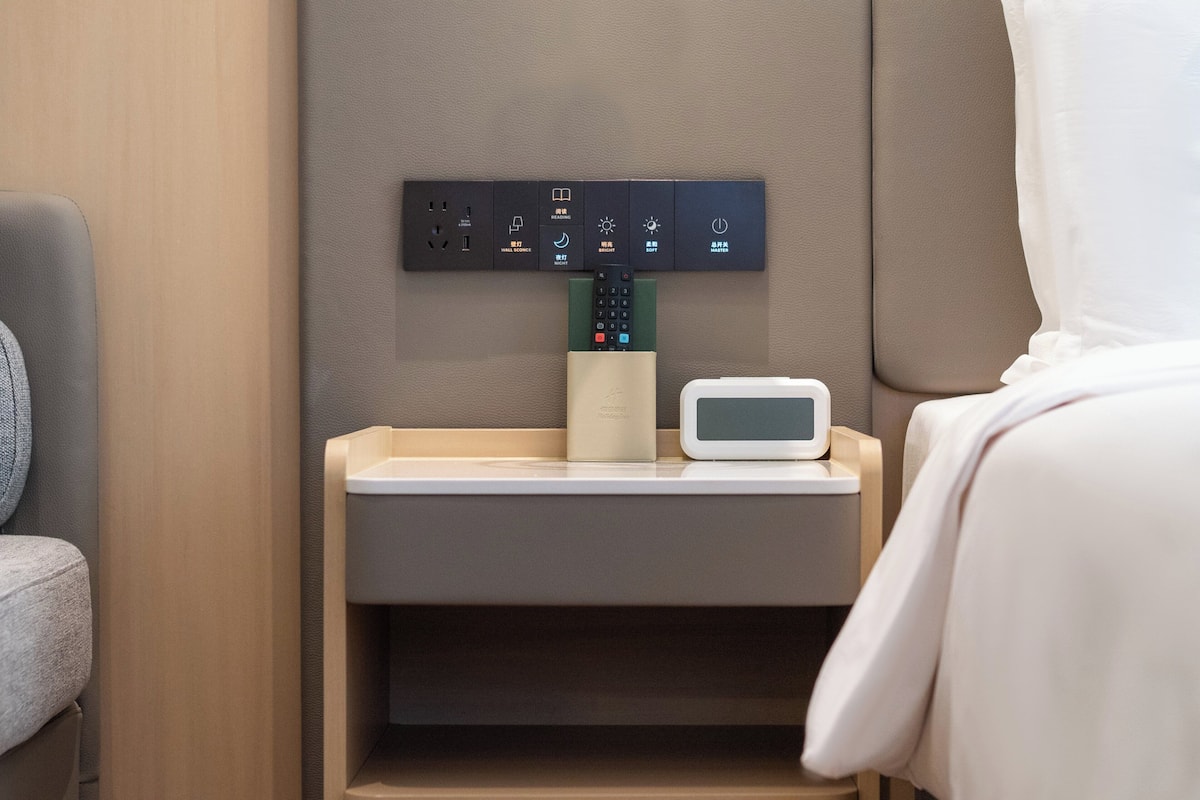 Ein Hotelzimmer mit Nachttisch, Bedienfeld mit verschiedenen Tasten, Fernbedienung und digitaler Uhr.