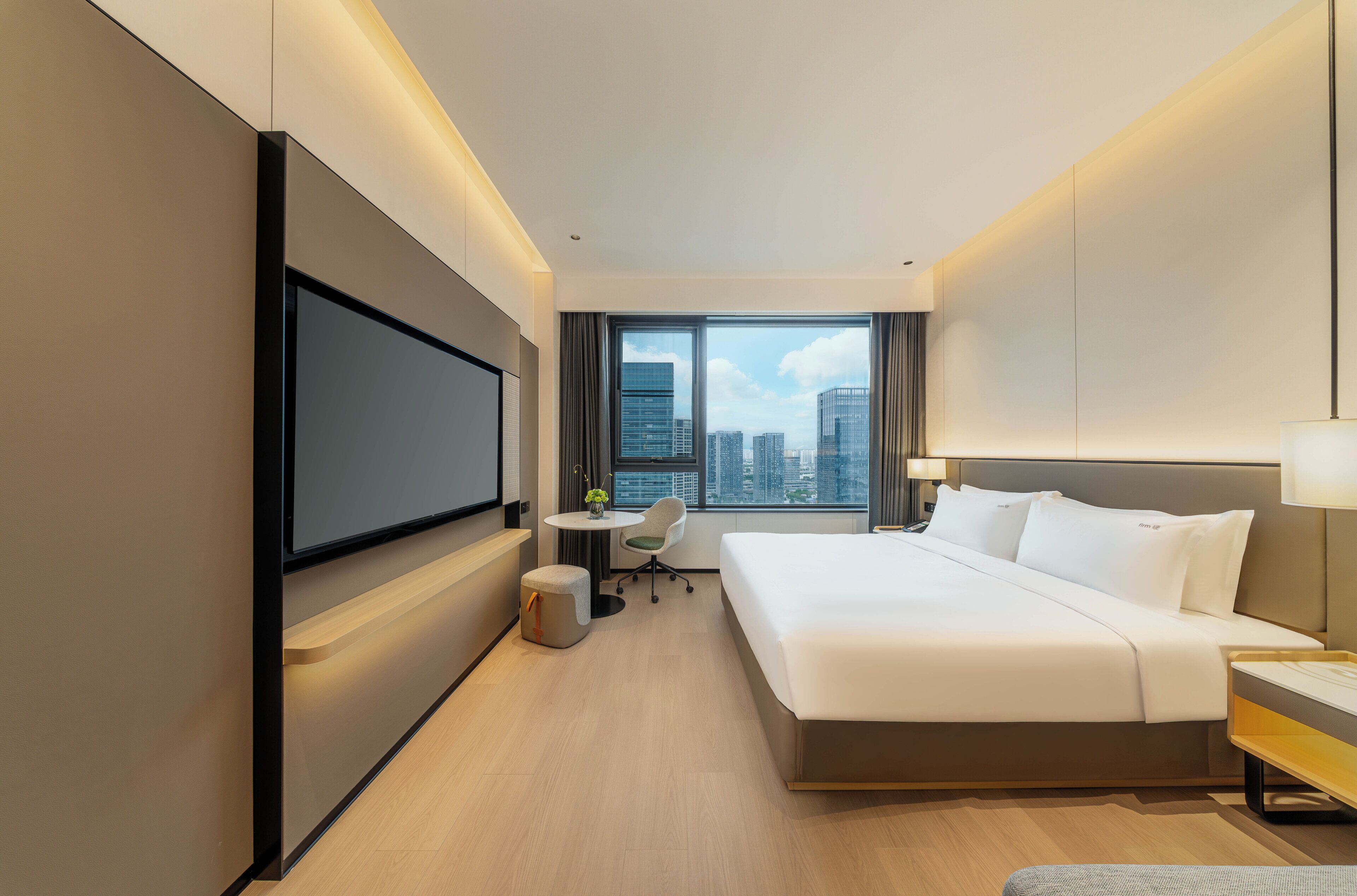 A modern hotel room with a large bed, a flat-screen TV mounted on the wall, a small desk with a chair, and a window offering a city view.