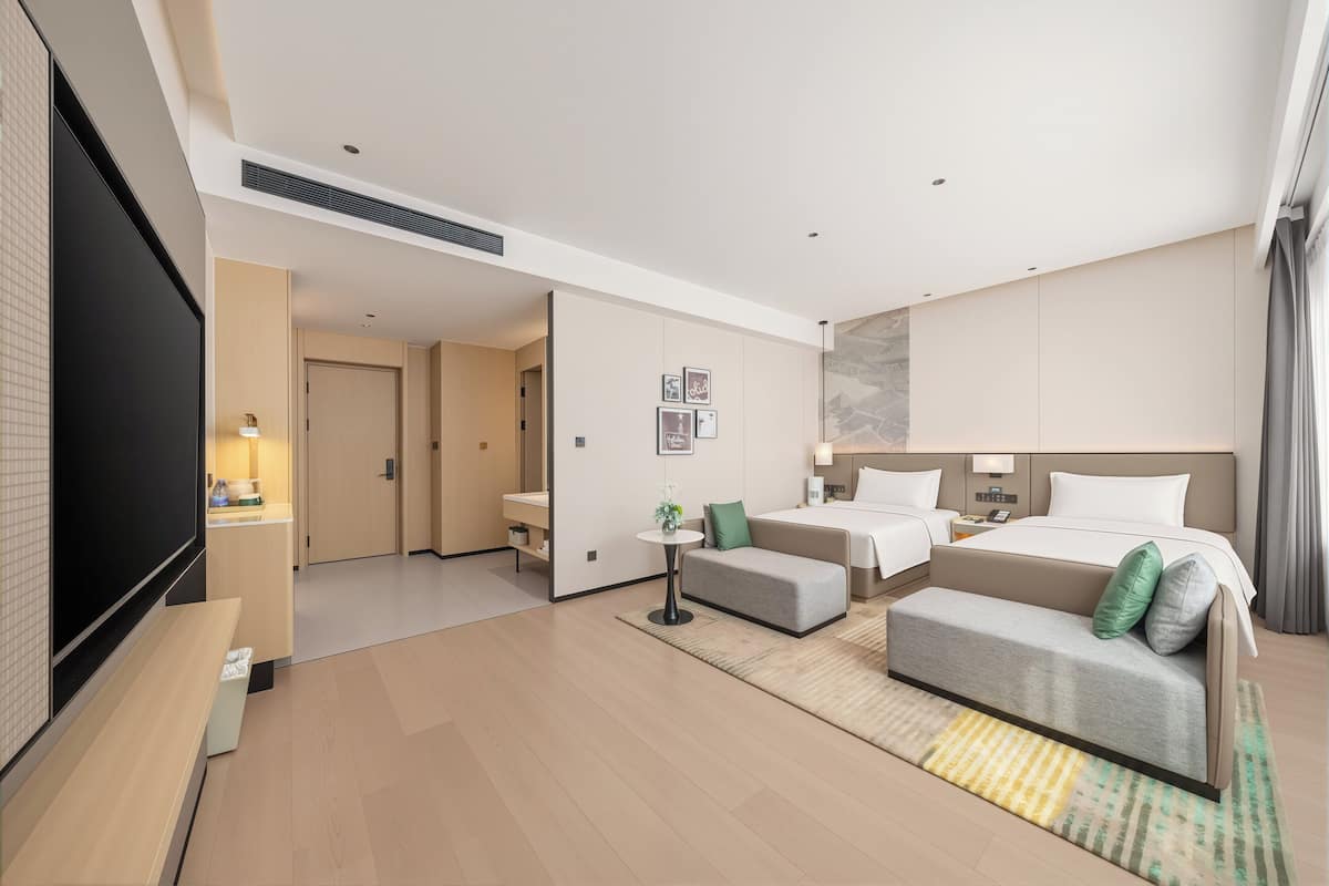 Ein modernes Hotelzimmer mit Flachbildfernseher, einer Couch, einem Bett, einem kleinen Tisch mit Pflanze und einem großen Fenster.