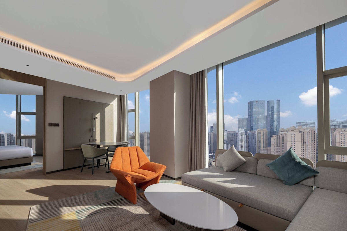 Ett modernt hotellrum med ett stort fönster som erbjuder utsikt över staden, en soffa, en orange fåtölj och en matplats.