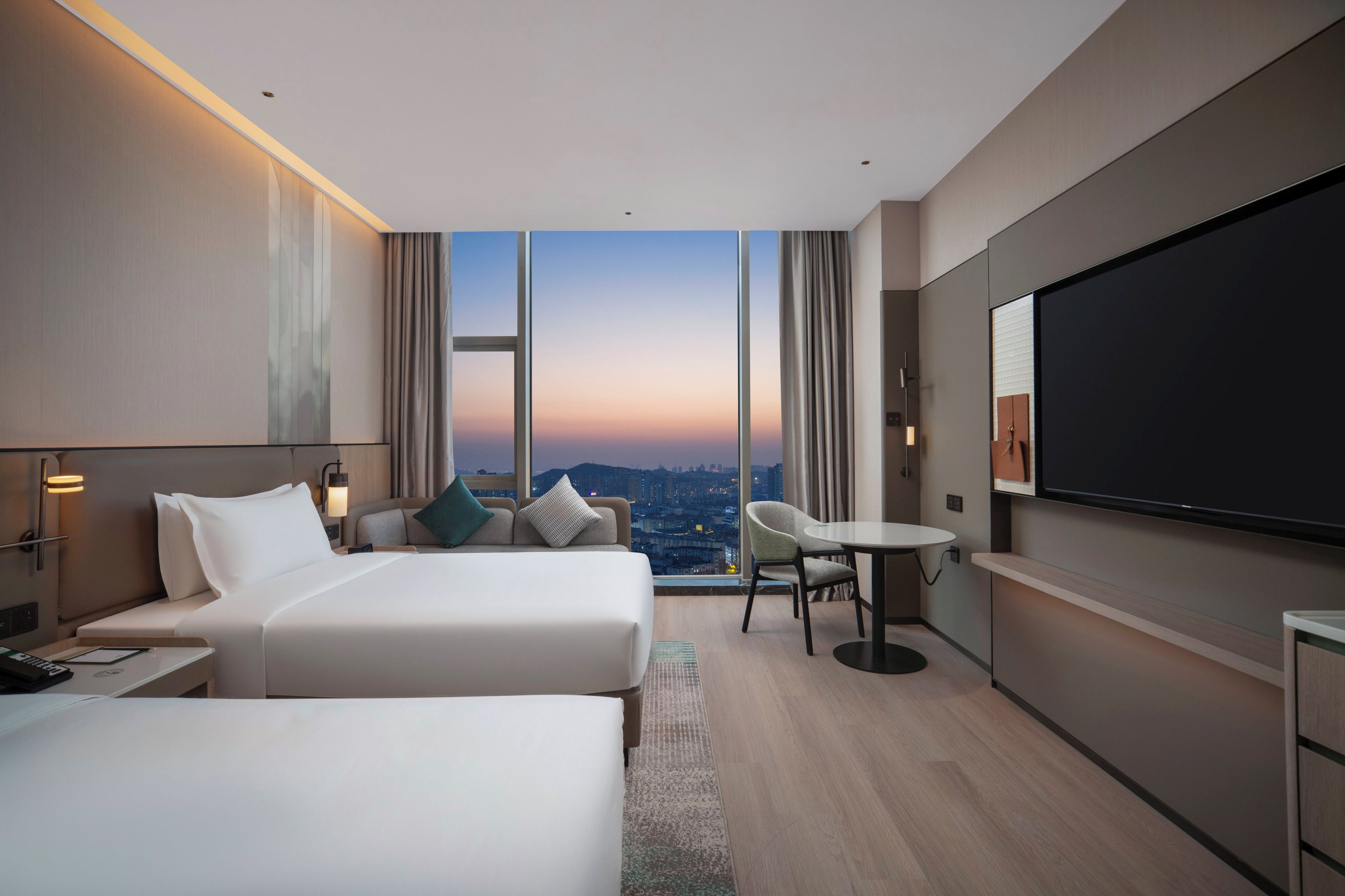 A hotel room with two beds, a desk, a chair, a TV, and a large window offering a city view.