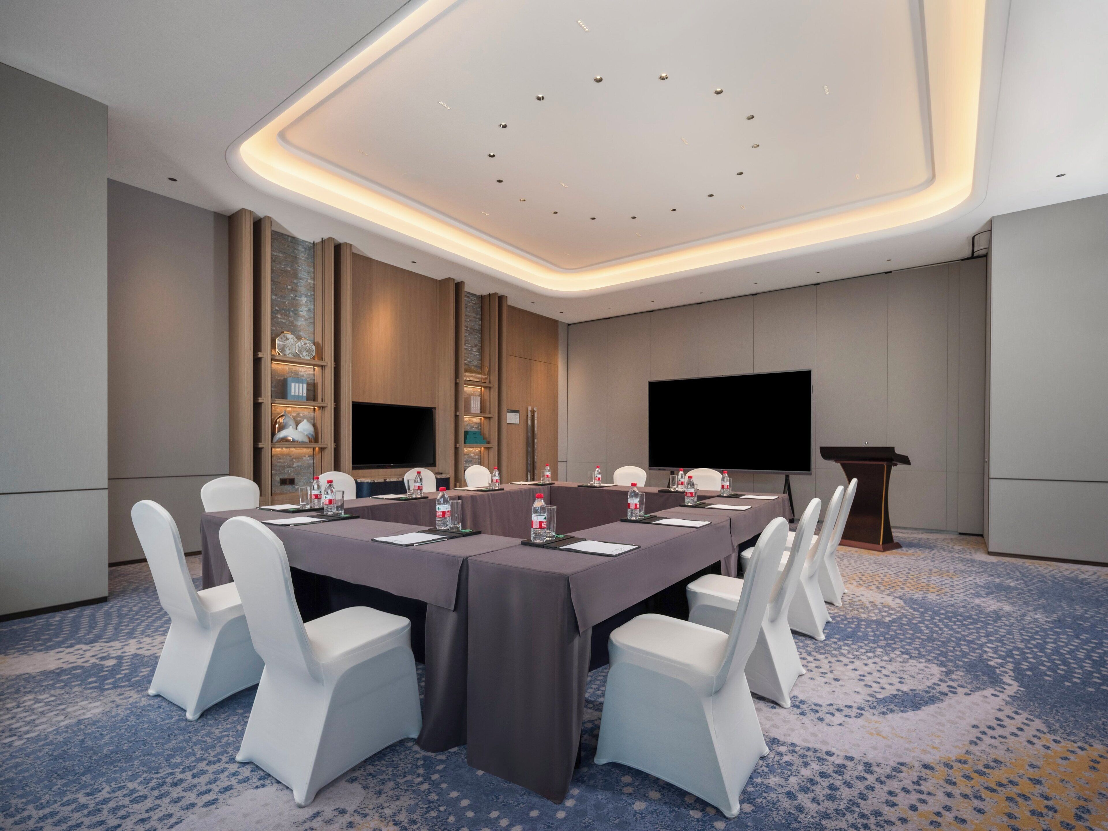 Une salle de conférence avec une longue table, des chaises blanches, un pupitre et un grand écran.