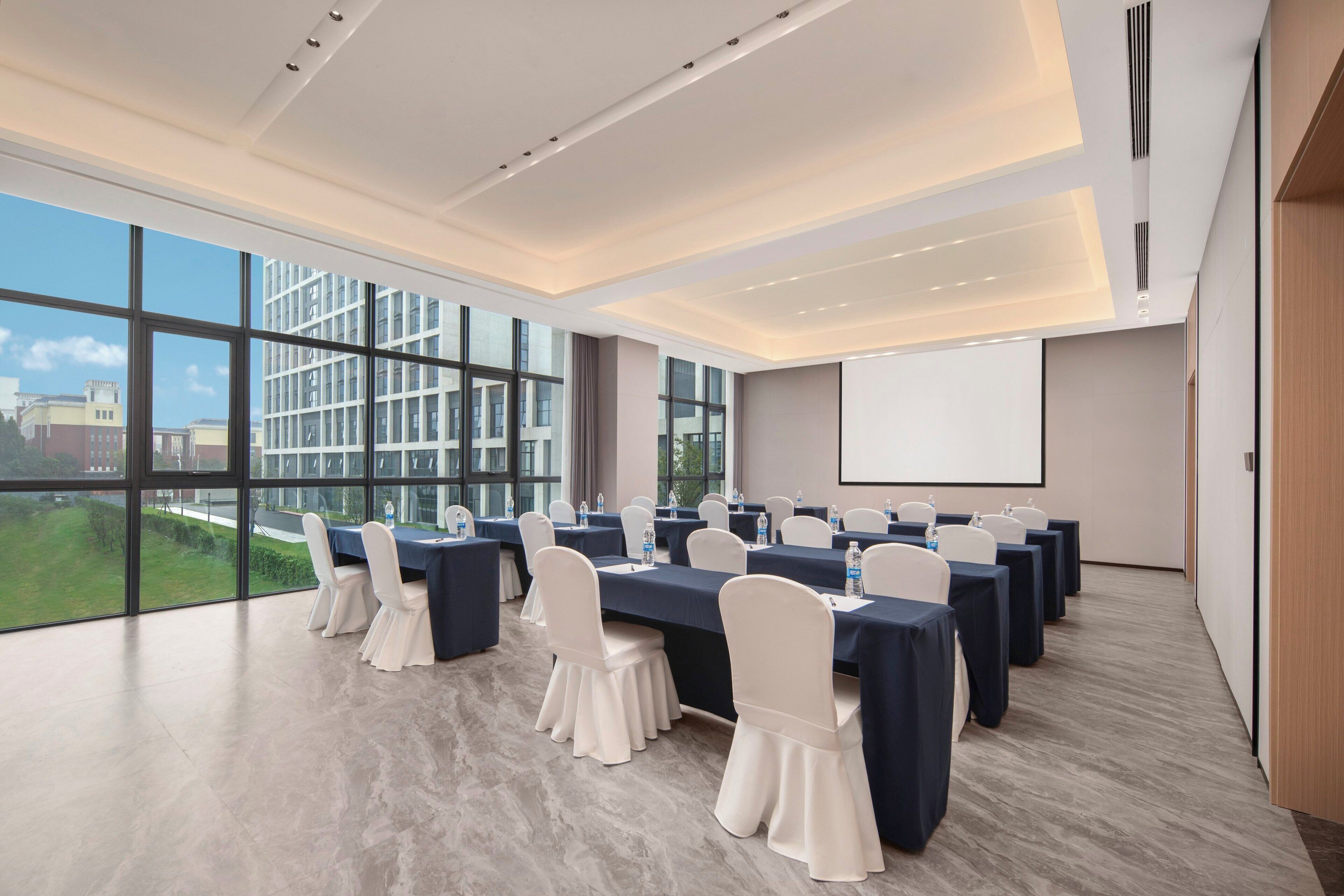 Una sala de conferencias con una gran mesa, sillas, una pantalla de proyector y vistas a edificios a través de amplios ventanales.