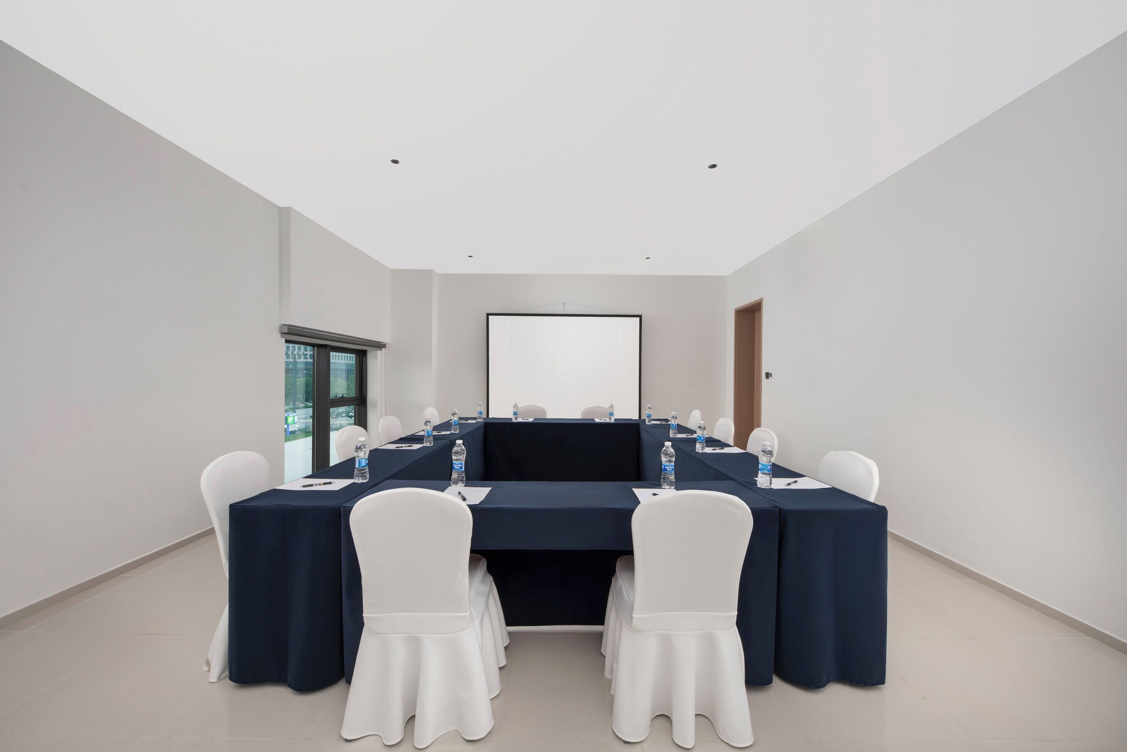 A conference room with a long table, white chairs, and a projector screen.