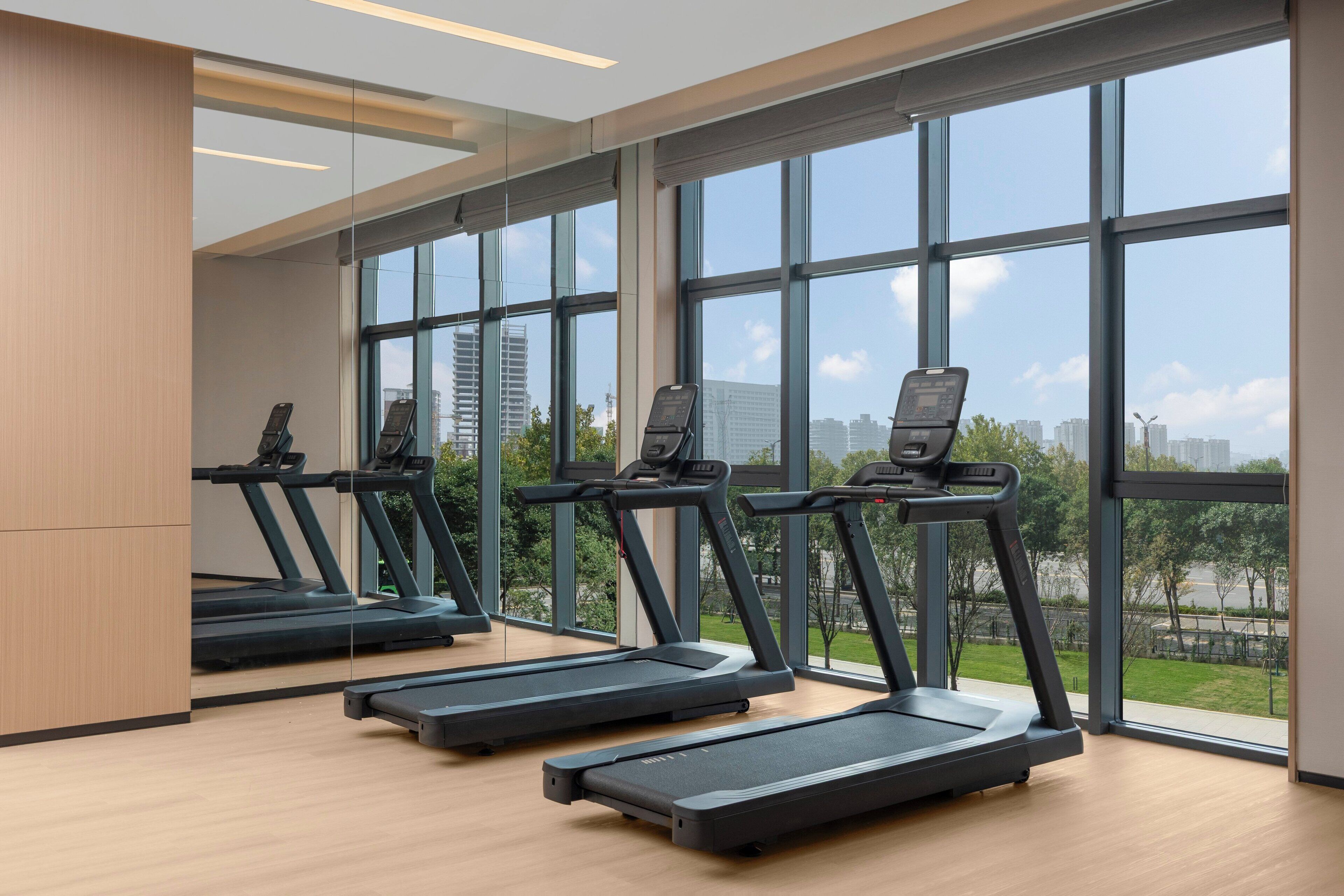A modern gym with four treadmills aligned in a row, large windows offering views of a cityscape and greenery.