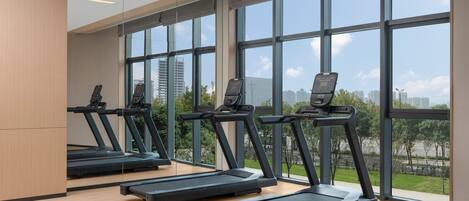 A modern gym with four treadmills aligned in a row, large windows offering views of a cityscape and greenery.