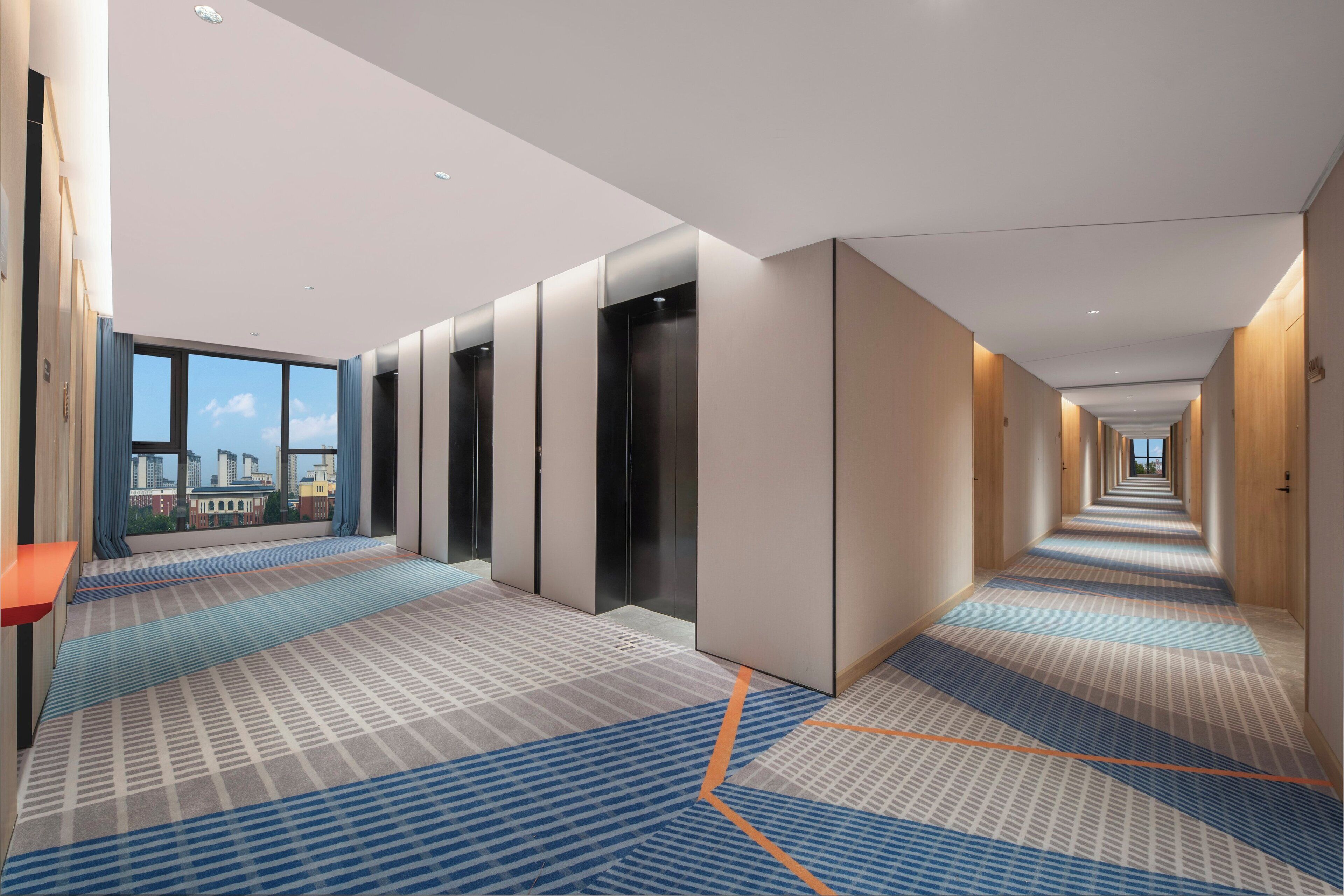 A modern hotel corridor with a patterned carpet, wooden doors, and a view of the city through large windows.