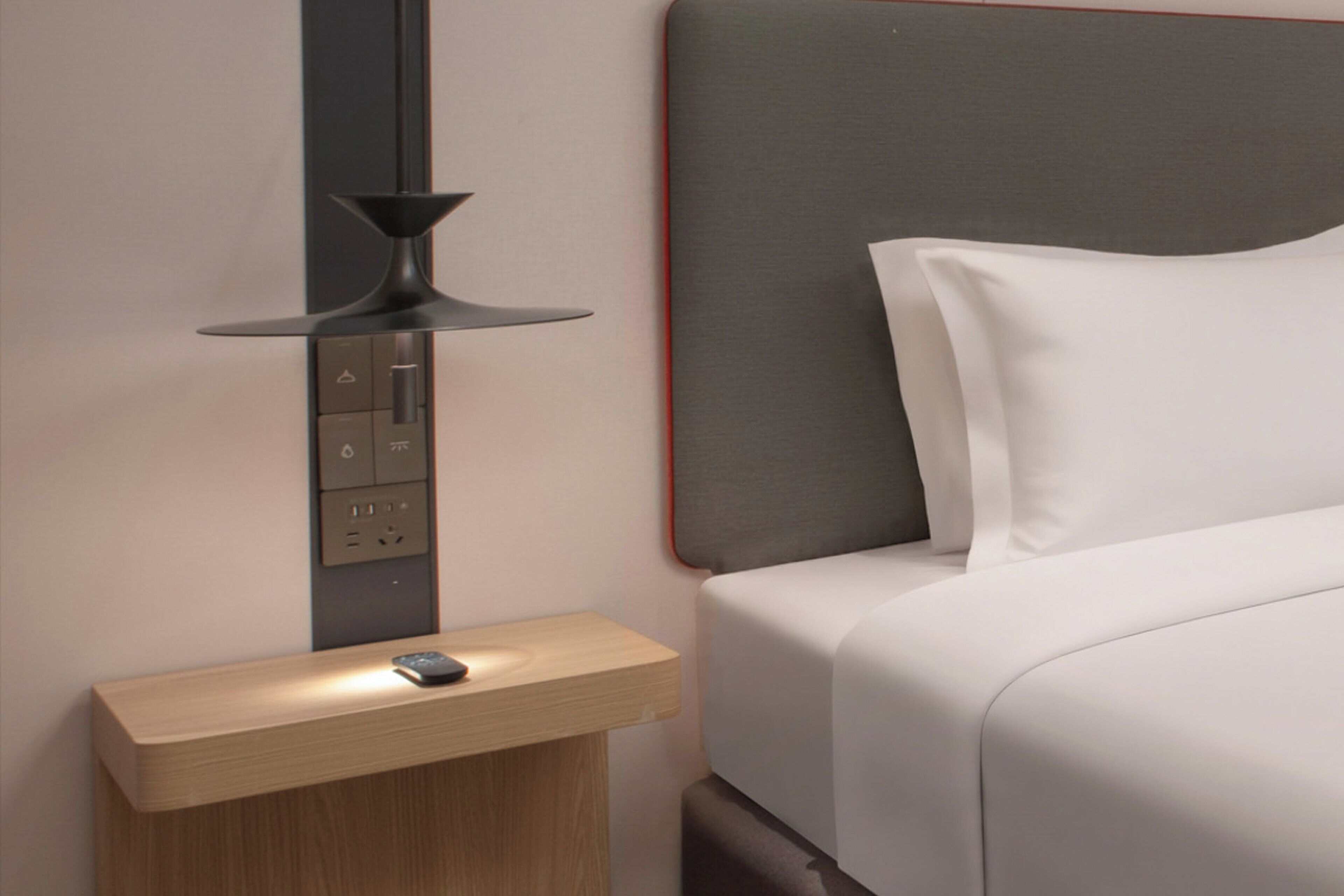 Moderni hotellihuone, jossa on puinen yöpöytä, seinään kiinnitetty valaisin ja siististi pedattu sänky.