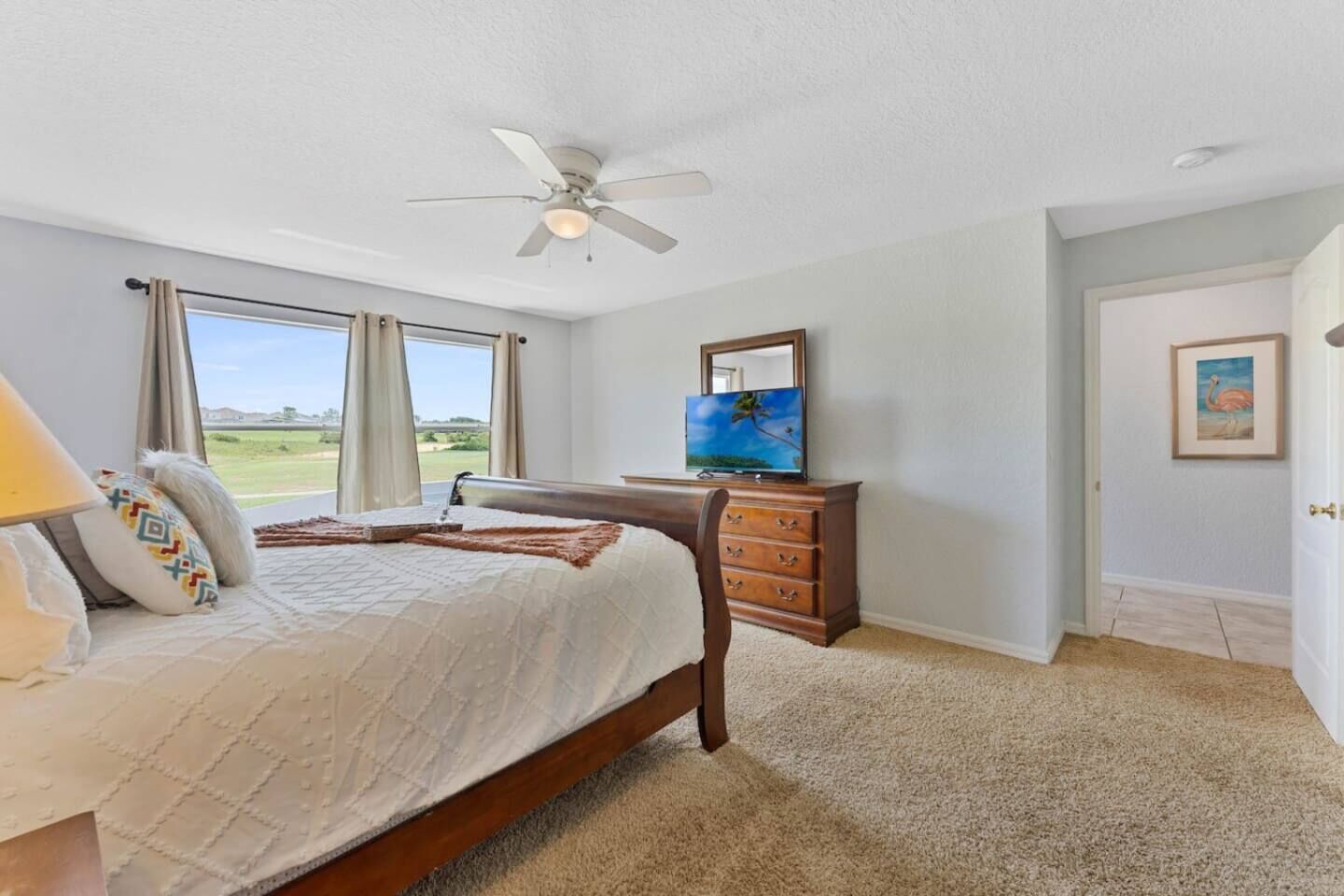 Quarto com cama, penteadeira, ventoinha no teto, televisão e janela com cortinas.