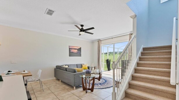 A living room with a sofa, a dining table, a ceiling fan, and a view of the outdoors through a glass door.