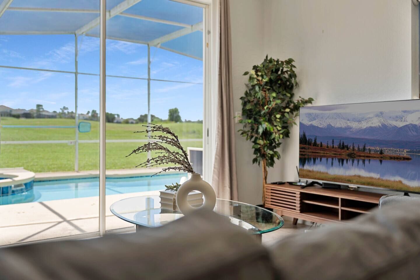 A living room with a glass table, a plant, a painting of a landscape, and a view of a swimming pool and green field.