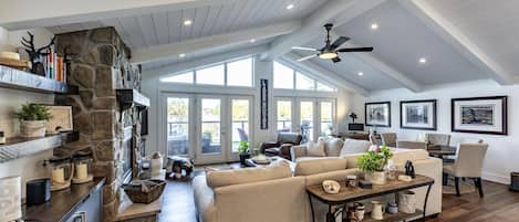 A spacious living room with a stone fireplace, a ceiling fan, and a large window overlooking a deck.