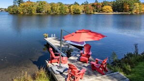 A dock with red Adirondack chairs and a red umbrella, overlooking a calm lake with autumn-colored trees.