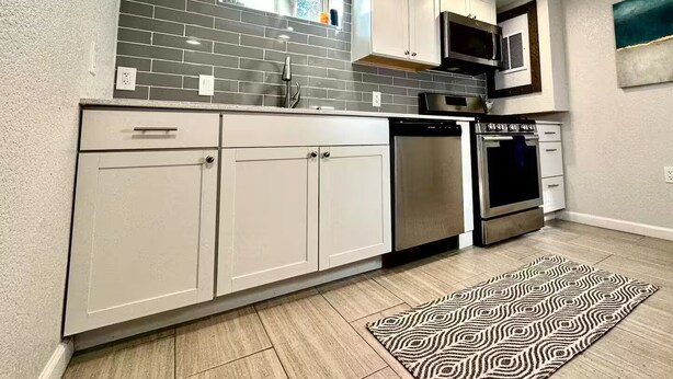 A modern kitchen with white cabinets, a grey backsplash, and stainless steel appliances. - The Cozy Below (Edgewater)