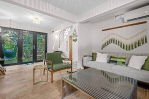 A modern living room with a glass coffee table, a sofa with cushions, a green armchair, and a hammock outside a sliding glass door.