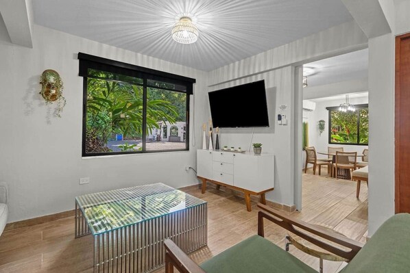 Un salon moderne avec un téléviseur, une table en verre, un meuble blanc et une vue sur la verdure à l’extérieur.