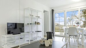 A modern living room with a flat-screen TV, white shelving units, a coffee table, and a dining area with a table and chairs.