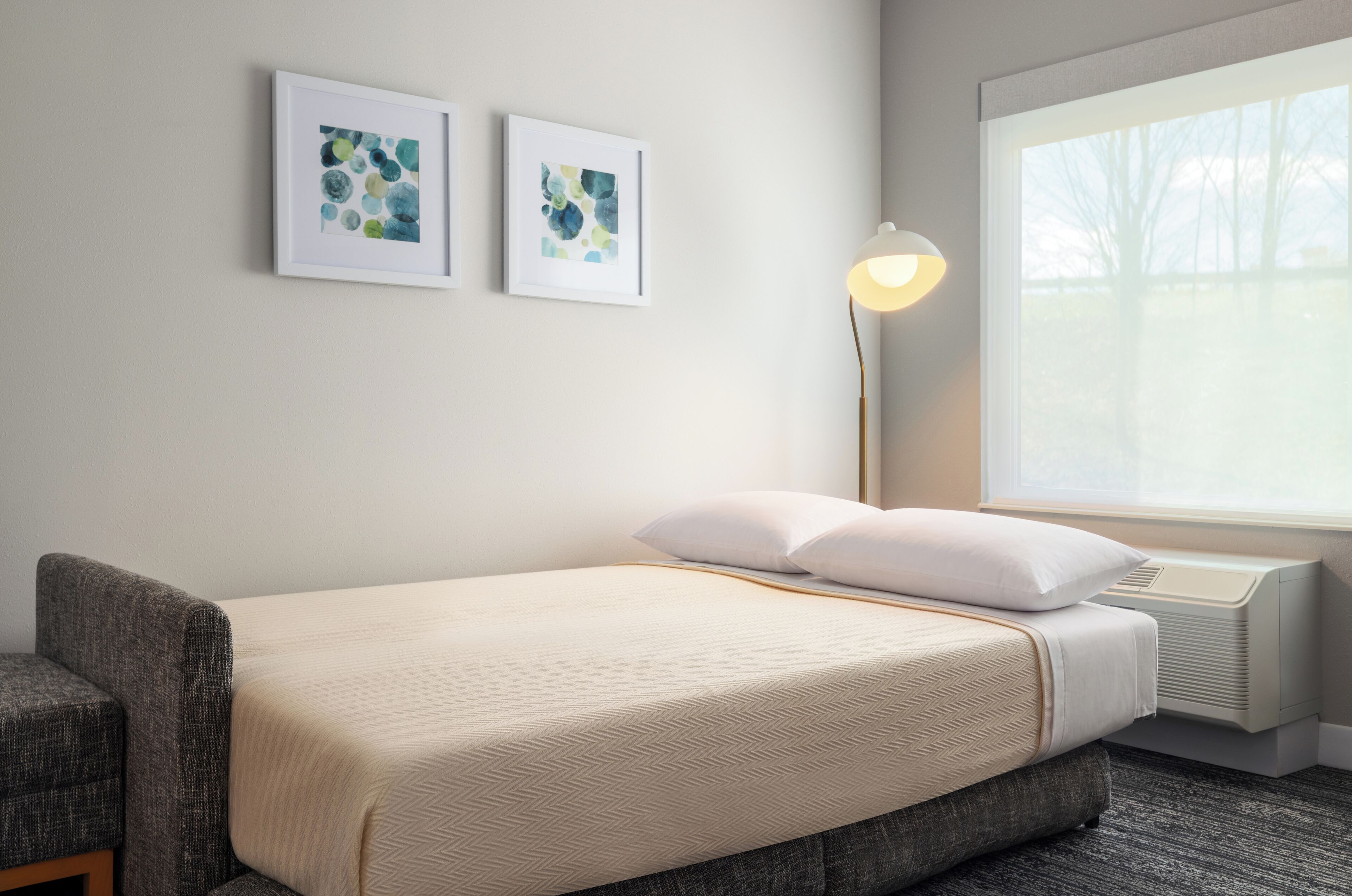 A hotel room with a bed, a chair, a standing lamp, and two framed artworks on the wall.
