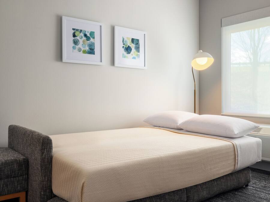 Een hotelkamer met een bed, een stoel, een staande lamp en twee ingelijste kunstwerken aan de muur.